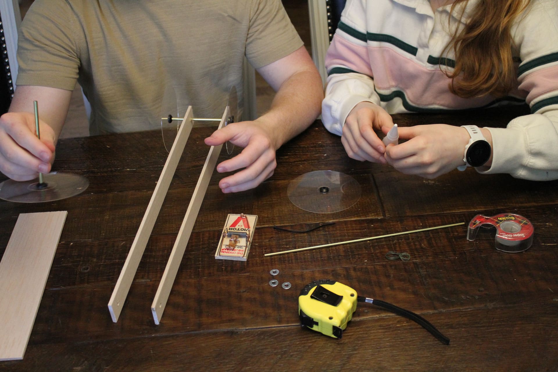 Two students mid-build, parts and tape measure laid out