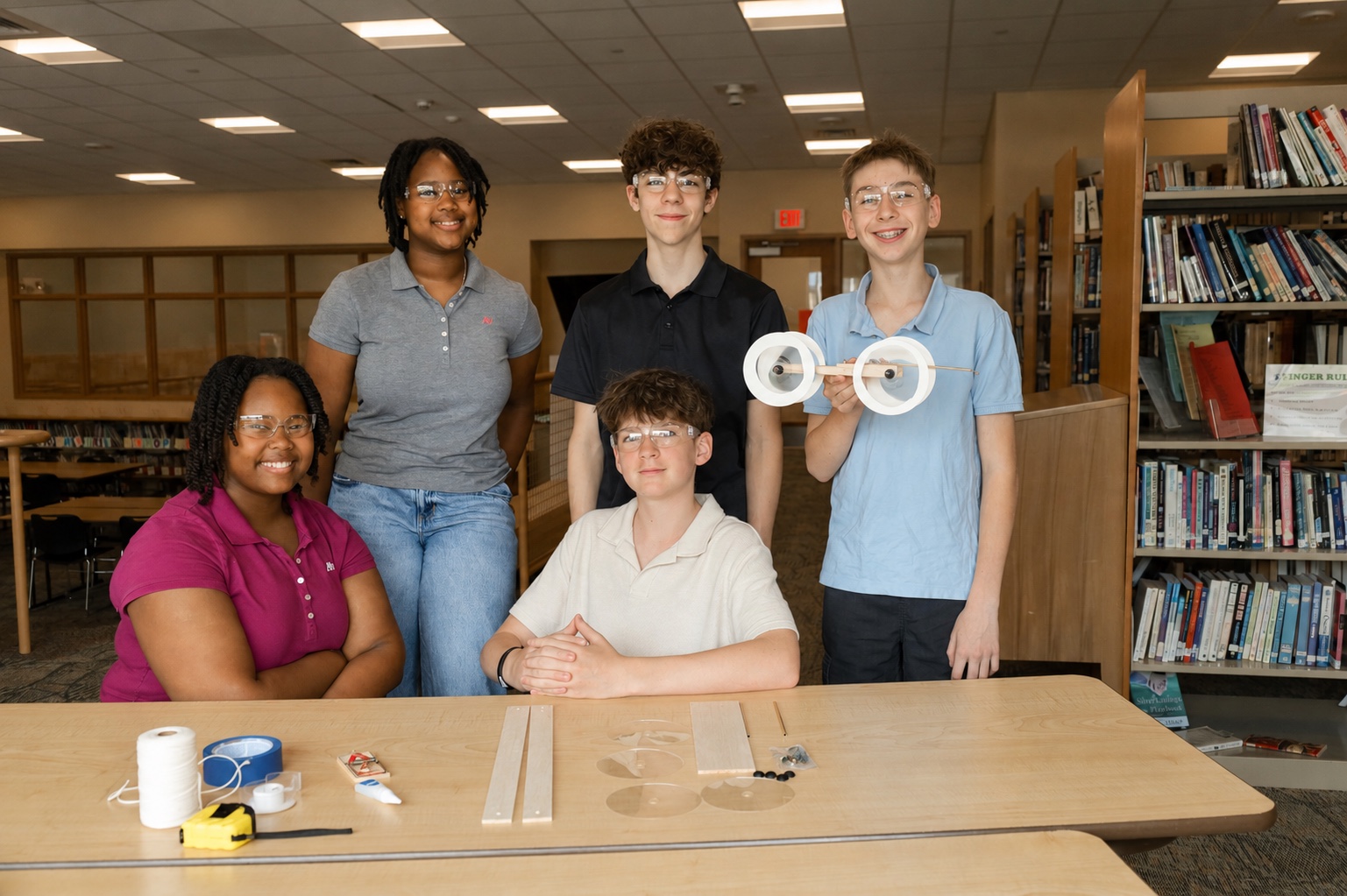 A group of kids working on the build together at a co-op table, several students and one adult, kits and mousetrap cars in frame.
